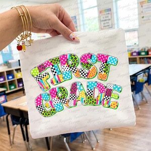 May include: A white t-shirt featuring the text FIRST GRADE in large, multi-colored letters. Each letter is filled with various patterns including leopard print, polka dots, stars, and checkerboard designs in bright neon colors like pink, green, and blue.