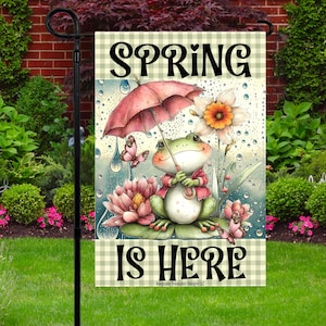Puede incluir: Bandera de jardín con una ilustración de una rana con un paraguas, rodeada de flores y mariposas. El texto "SPRING IS HERE" está presente. La bandera tiene un borde a cuadros y está diseñada para uso en exteriores.