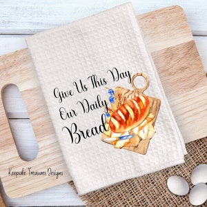 May include: White kitchen towel with the text "Give Us This Day Our Daily Bread" in black cursive font. A watercolor illustration of a loaf of bread on a wooden cutting board with a knife and a sprig of blue flowers.