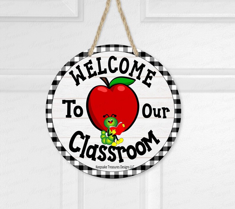 May include: Round wooden sign with a black and white checkered border. The sign reads "WELCOME To Our Classroom" with a red apple and a cartoon caterpillar holding a book. A jute rope is attached for hanging.