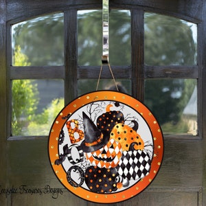 May include: A round Halloween door hanger with a white background and orange, black, and white pumpkins. The pumpkins have black polka dots and a witch's hat. The text "BOO" is written in black and white letters.