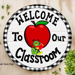 A round welcome sign with a black and white checkered border. The sign has a white background with black text that reads "Welcome to our Classroom". A red apple with a green stem and leaf is in the center of the sign. A green worm with a smiling face is sitting on the apple and reading a book.