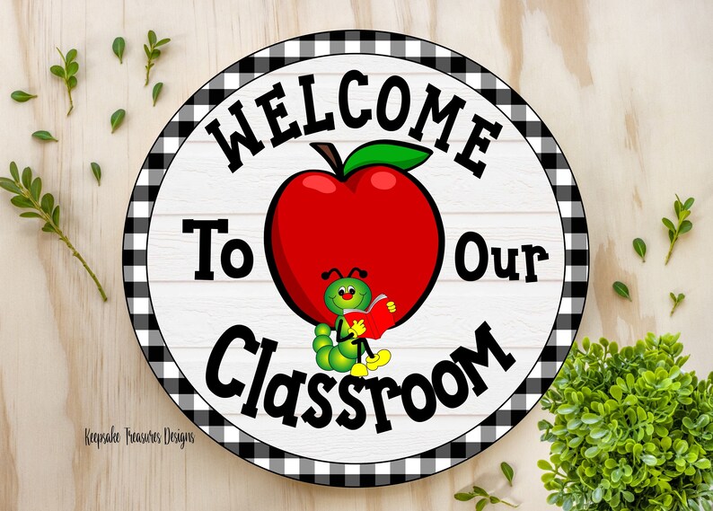 A round welcome sign with a black and white checkered border. The sign has a white background with black text that reads "Welcome to our Classroom". A red apple with a green stem and leaf is in the center of the sign. A green worm with a smiling face is sitting on the apple and reading a book.
