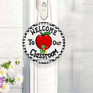 May include: Round wooden sign with the text "WELCOME To Our Classroom" in black lettering. A red apple with a green leaf and a cartoon worm holding a book are in the center. The sign has a black and white checkered border and is hanging on a white door.