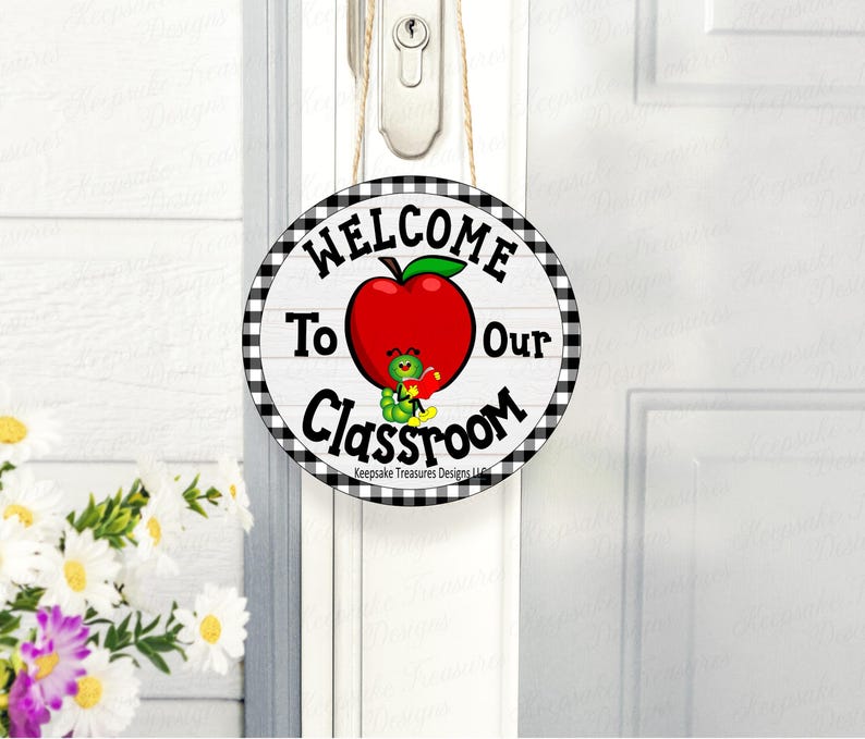 May include: Round wooden sign with the text "WELCOME To Our Classroom" in black lettering. A red apple with a green leaf and a cartoon worm holding a book are in the center. The sign has a black and white checkered border and is hanging on a white door.