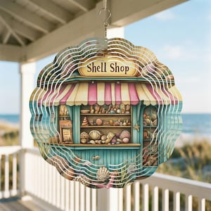Pode incluir: Um spinner de vento decorativo de metal em camadas com uma cena caprichosa de "Shell Shop". A loja tem um toldo listrado rosa e branco, prateleiras cheias de conchas e o texto "Shell Shop" acima da entrada. O fundo mostra uma praia e o oceano.