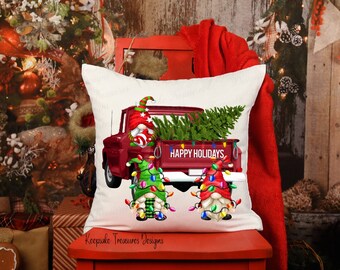 May include: White throw pillow with a red truck carrying a Christmas tree and two gnomes decorated with Christmas lights. The truck has the words "Happy Holidays" on the back.