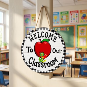 May include: Round wooden classroom sign with a black and white checkered border. The sign features the words "WELCOME To Our Classroom" around a red apple with a green leaf and a cartoon caterpillar holding a book. A rope is attached for hanging.