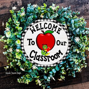 Welcome to Our Classroom sign with a black and white checkered border. The sign features a red apple with a smiling green worm holding a book. The sign is surrounded by a green wreath with white flowers.