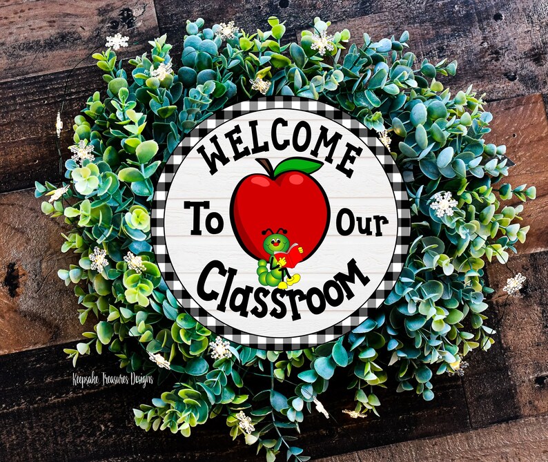 Welcome to Our Classroom sign with a black and white checkered border. The sign features a red apple with a smiling green worm holding a book. The sign is surrounded by a green wreath with white flowers.
