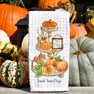 May include: A white kitchen towel with a watercolor illustration of a tiered tray decorated with pumpkins, gourds, leaves, and a pie. The tray has a sign that says "Hello Fall". The towel has the text "Keepsake Treasures Designs" at the bottom.