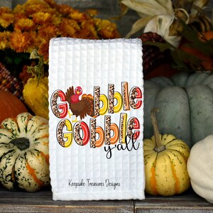 May include: White kitchen towel with the text "Gobble Gobble y'all" in orange, yellow, and brown letters with a leopard print design. A small turkey illustration is in the top left corner of the text.