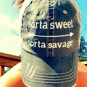 May include: A gray and white baseball cap with a mesh back. The cap has the text "sorta sweet sorta savage" embroidered on the front with an arrow pointing to the right.