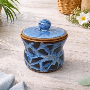 May include: A ceramic jar with a lid, featuring a blue and black glaze design. The jar has a rounded shape with a lid and a small knob on top. The jar is sitting on a wooden surface.
