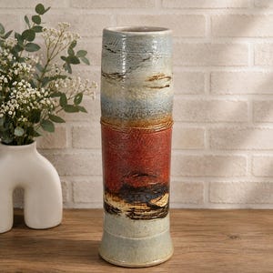 May include: A tall, cylindrical ceramic vase featuring a multi-coloured glaze. The top section is a pale blue-grey, the middle is a deep rust-red, and the base is a light cream colour. The surface has a textured, layered appearance with dark brown accents.