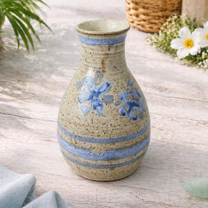 May include: A beige ceramic vase with blue floral designs and horizontal blue stripes. The vase has a narrow neck and a rounded body. The vase is likely used for decorative purposes or holding flowers.
