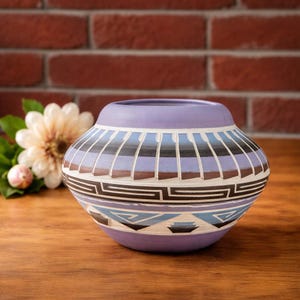 May include: A hand-painted ceramic pot with a rounded shape and a wide opening. The pot features a lavender base and rim, with geometric patterns in blue, black, and brown. The design includes vertical stripes and angular shapes. The pot is displayed on a wooden surface.
