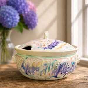 May include: A round ceramic lidded tureen featuring a swirling, multi-colored glaze in shades of purple, blue, pink, and cream. The bowl has two small side handles and a matching lid with a central knob, showcasing a glossy, textured finish.