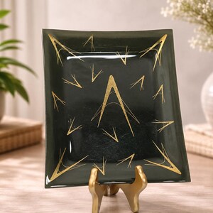 Puede incluir: Un plato cuadrado de cristal negro con una letra A dorada en el centro y detalles dorados en forma de V dispersos. El plato se exhibe en posición vertical sobre un soporte de metal dorado, mostrando un acabado brillante y un patrón geométrico.