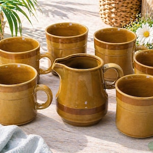 May include: A collection of six ceramic mugs and a jug in shades of brown. The mugs are cylindrical with handles and a darker brown band. The jug has a spout and handle, with a similar band. The set is arranged on a light-coloured surface, with a natural theme.
