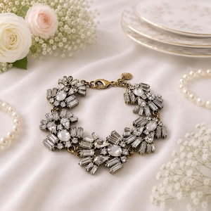 Op de afbeelding: Een vintage-stijl armband met heldere, sprankelende kristallen en een goudkleurige metalen setting. De armband heeft een bloemenontwerp met een sluiting. De sieraden worden gepresenteerd op een witte satijnen ondergrond, met bloemen en parels op de achtergrond.