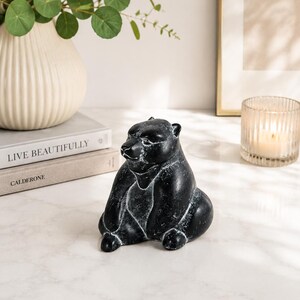 Könnte beinhalten: Eine schwarze Bärenfigur mit einem abgenutzten, verwitterten Finish und sichtbaren weißen Akzenten entlang der Konturen. Der Bär ist in sitzender Position dargestellt, wobei die Vorderpfoten auf dem Boden ruhen und eine glatte Form zeigen.