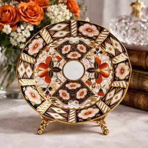 May include: A decorative porcelain plate with a floral design in orange, white, and navy blue, accented with gold trim. The plate is displayed on a gold-colored stand, showcasing its intricate details and craftsmanship.