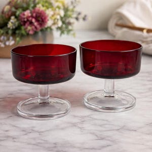 May include: Two ruby red glass dessert bowls with a clear stem and base. The bowls have a wide, shallow design. The stem and base are clear glass. The background includes a vase of flowers and a white cloth.