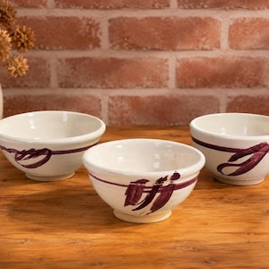 May include: Three off-white ceramic bowls with a glossy finish, each featuring a hand-painted dark purple horizontal stripe and abstract brushstroke accents around the exterior. The bowls have a rounded shape with a small circular base.