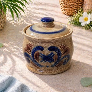 May include: A tan ceramic jar with a lid, featuring blue and brown floral designs. The jar has a textured surface and a blue knob on top. The lid has a blue stripe. The jar is likely for kitchen or home decor.