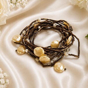 May include: A necklace featuring large, flat, cream-coloured beads and smaller white beads, strung on a dark brown cord. The necklace is arranged in a spiral on a white satin surface, with white flowers in the background.