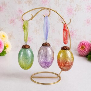 May include: Three oval-shaped glass ornaments with a crackled texture, hanging from a gold-toned metal stand. The ornaments are green, purple, and a yellow-to-red gradient, each topped with a decorative bronze-coloured cap and a coloured ribbon.