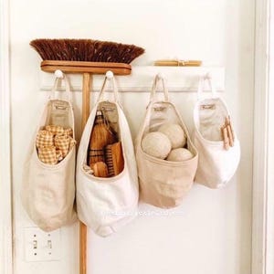May include: Four beige canvas bags hanging on a white wall with a wooden broom and a wooden shelf above. The bags are filled with various items, including cleaning supplies, laundry balls, and cloths.