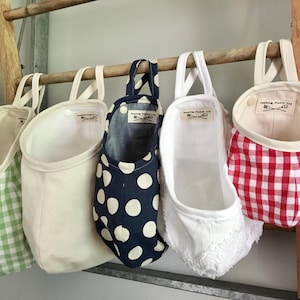 May include: Five hanging fabric storage pouches with loops, featuring various patterns including green gingham, solid cream, navy blue with white polka dots, white textured fabric, and red gingham. Each pouch has a label reading Feeding Pickle Ltd.