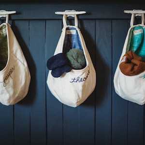 May include: Three off-white canvas hanging storage pouches suspended from white cleats. Each pouch features embroidered names: jasper, theodore, and lillie. The interiors are lined with green, blue, and teal fabric, each holding a folded knit hat.