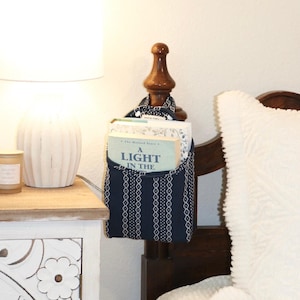 May include: A navy blue and white patterned bedside organizer hanging on a wooden bed frame. The organizer holds books, including one titled "A Light in the". A white lamp and a candle sit on a nearby nightstand.