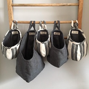 May include: Five hanging storage baskets in varying sizes. The baskets are made of gray fabric and white fabric with black vertical stripe patterns. They hang from a wooden ladder. Each basket has a small tag.