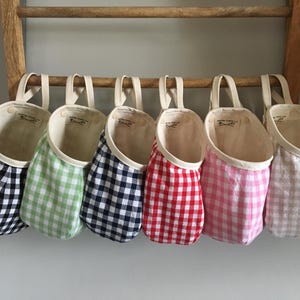 May include: Six fabric storage baskets in a row, each with a different color gingham pattern: black, green, navy, red, pink, and tan. The baskets hang from a wooden ladder, with cream-colored handles and interior lining.
