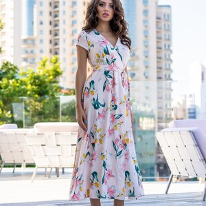 May include: A woman wearing a white maxi dress with a floral print. The dress has a V-neckline, short sleeves, and a tie at the waist. She is standing on a wooden deck.