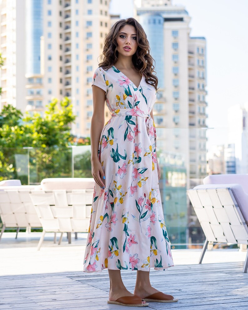 May include: A woman wearing a white maxi dress with a floral print. The dress has a V-neckline, short sleeves, and a tie at the waist. She is standing on a wooden deck.