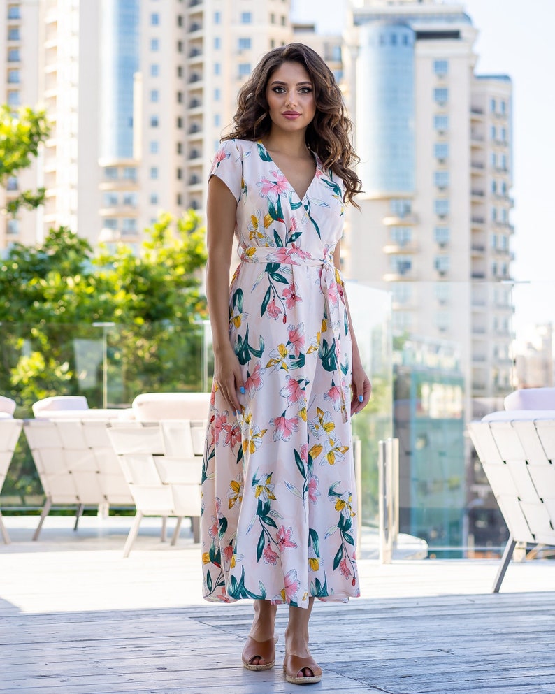 May include: A woman wearing a white maxi dress with a floral print. The dress has a V-neckline, short sleeves, and a tie at the waist. She is standing on a wooden deck with a city skyline in the background.