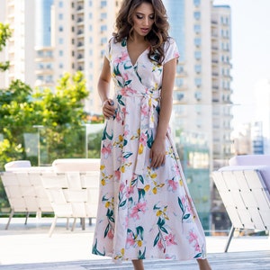 May include: A woman wearing a long, pink floral dress with a V-neckline and short sleeves. The dress has a tie at the waist and a flowing skirt. She is standing on a wooden deck with a city skyline in the background.