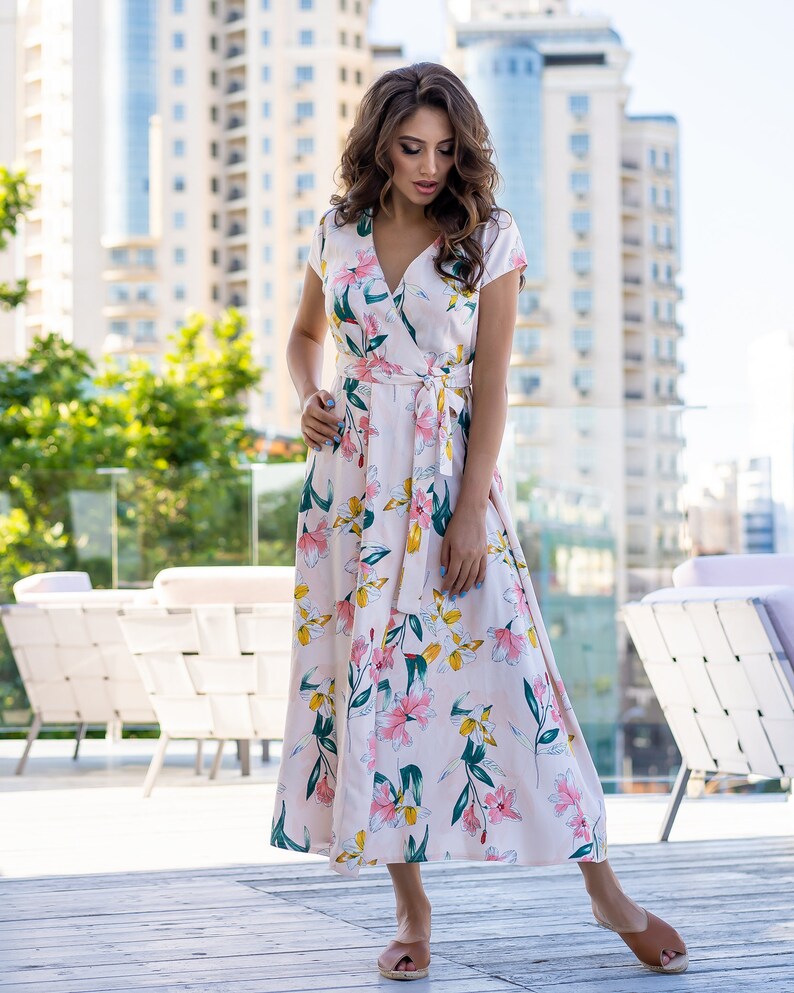 May include: A woman wearing a long, pink floral dress with a V-neckline and short sleeves. The dress has a tie at the waist and a flowing skirt. She is standing on a wooden deck with a city skyline in the background.