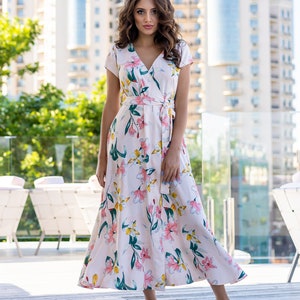 May include: A woman wearing a white maxi dress with a floral print. The dress has a V-neckline, short sleeves, and a tie at the waist. The woman is standing on a wooden deck.