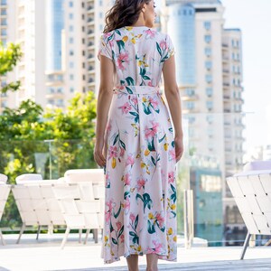May include: A woman wearing a long, pink floral dress with a white background. The dress has a V-neckline and short sleeves. The woman is standing in front of a building with a glass railing.