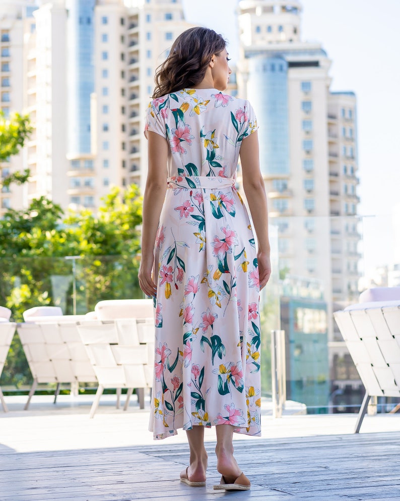 May include: A woman wearing a long, pink floral dress with a white background. The dress has a V-neckline and short sleeves. The woman is standing in front of a building with a glass railing.