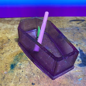 May include: A purple glitter coffin-shaped trinket dish with a pink and green pencil inside.