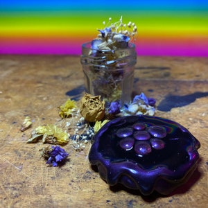 Puede incluir: Un adorno de resina de color morado oscuro con un diseño en relieve de un grupo de puntos redondos de color morado iridiscente. El adorno está sobre una superficie de madera con flores secas y un frasco de vidrio.