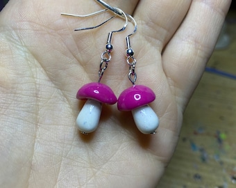 May include: A pair of pink and white mushroom earrings with silver hooks. The mushroom caps are a sparkly pink color and the stems are white with a glittery finish.