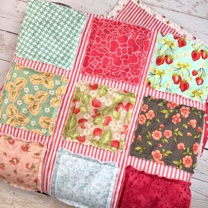 May include: A patchwork quilt with a variety of patterned squares, including floral, strawberry, and geometric designs. The squares are set against a red and white striped background. The quilt is made of soft fabric.